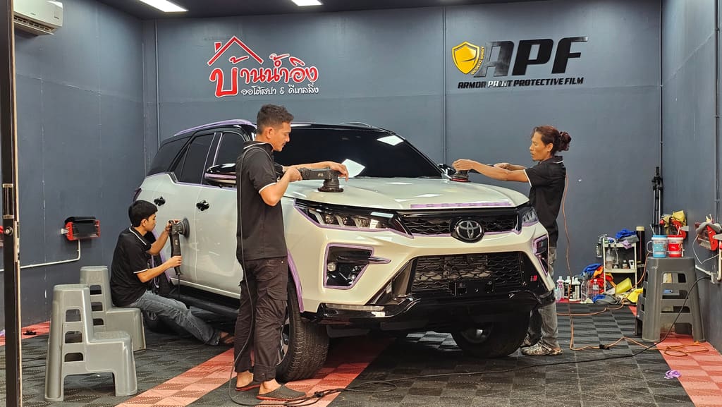 ช่างสามคนติดฟิล์มใสกันรอยให้ Toyota Fortuner สีขาวในห้องทำงานของร้าน เห็นป้ายบ้านน้ำอิงบนผนังด้านหลัง