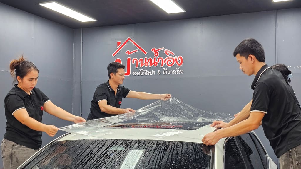 ช่างสามคนช่วยกันวางแผ่นฟิล์มใสกันรอยแผ่นใหญ่ลงบนรถ ด้านหลังเป็นโลโก้บ้านน้ำอิงบนผนังห้องทำงาน